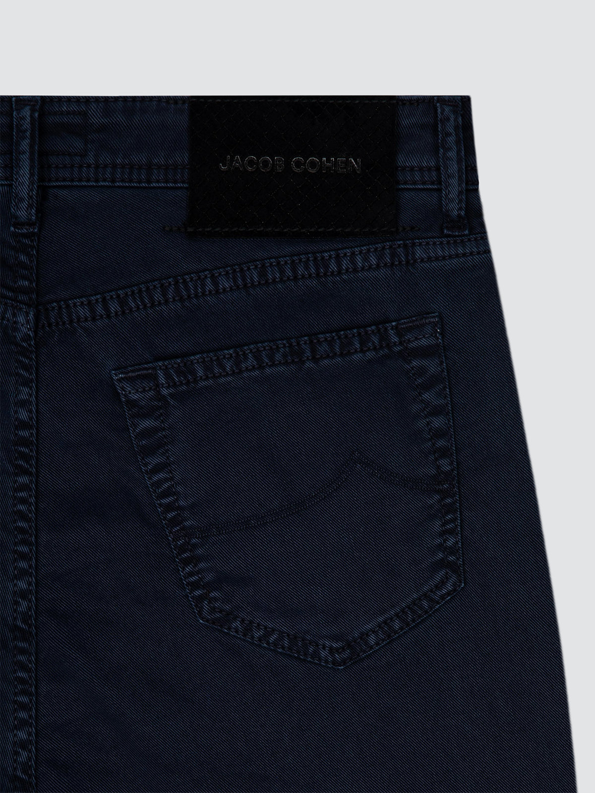 Close-up of the back waistband and pocket of dark navy denim jeans, showing a black fabric brand patch with 'JACOB COHEN' .
