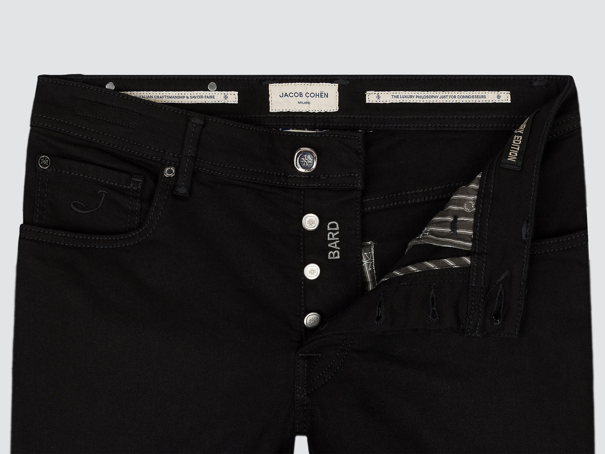 Close-up of black denim jeans showing the waistband, button fly, front pocket, and interior side pocket with a striped lining. Visible branding includes a central label reading JACOB COHEN and BARD on the fly.