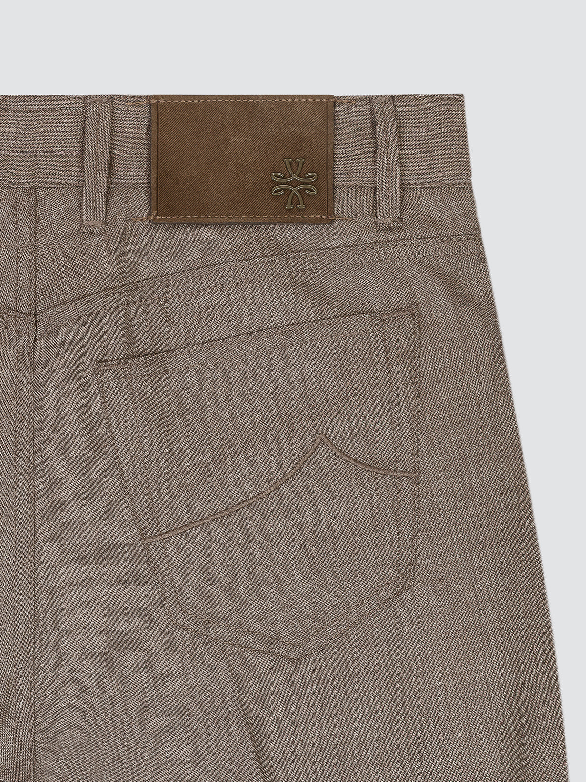 Close-up of the back waistband and pocket area of taupe-brown textured trousers, featuring a brown leather patch with an embossed logo on the back waist and a curved welt pocket.