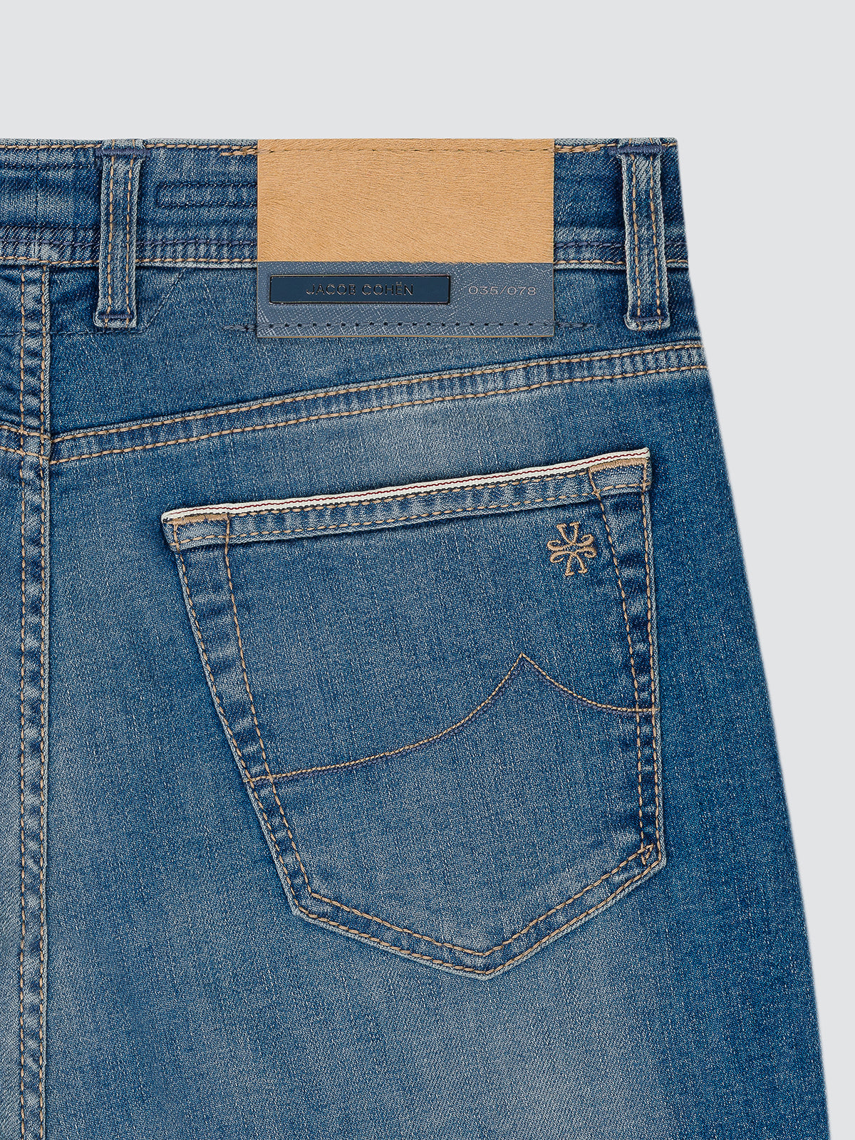Close-up of the back waistband and pocket area of blue denim jeans, showing a tan paper label, a dark blue brand patch, and a decorative embroidered logo on the pocket. jacob cohen ltd