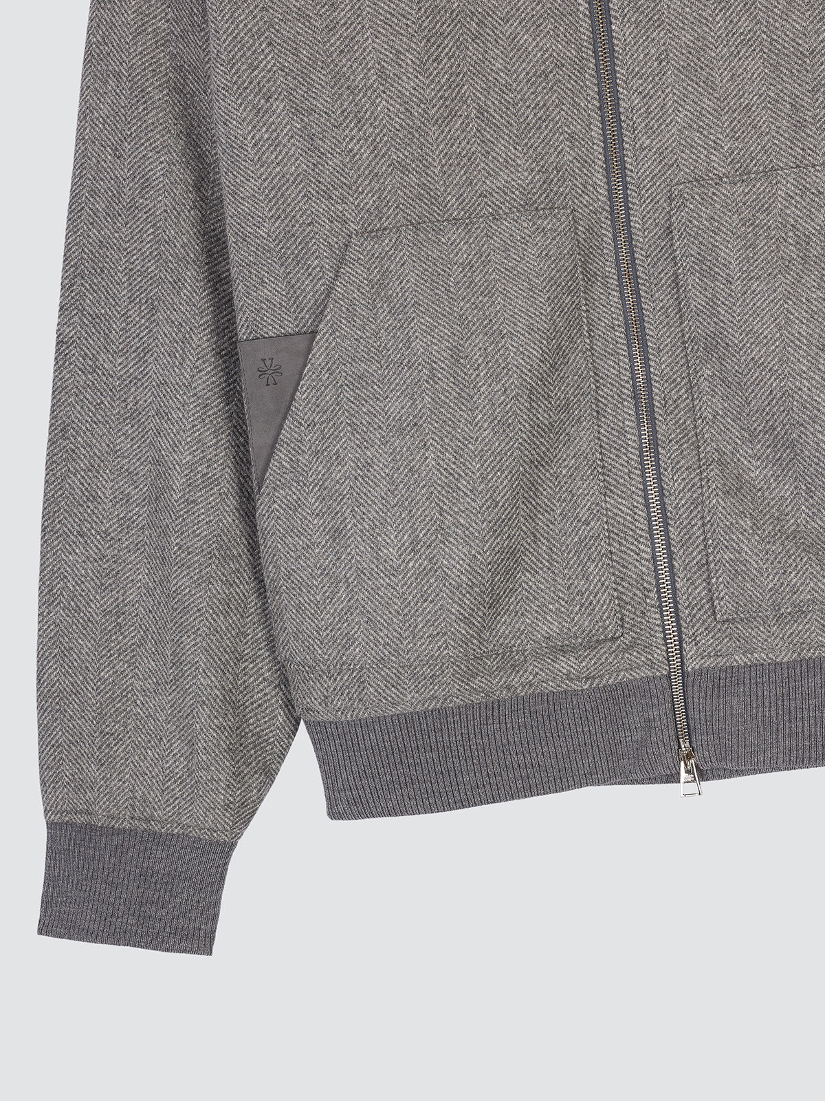 Close-up of a gray herringbone zip-up jacket with ribbed cuffs and hem, featuring a diagonal patch pocket on the sleeve.
