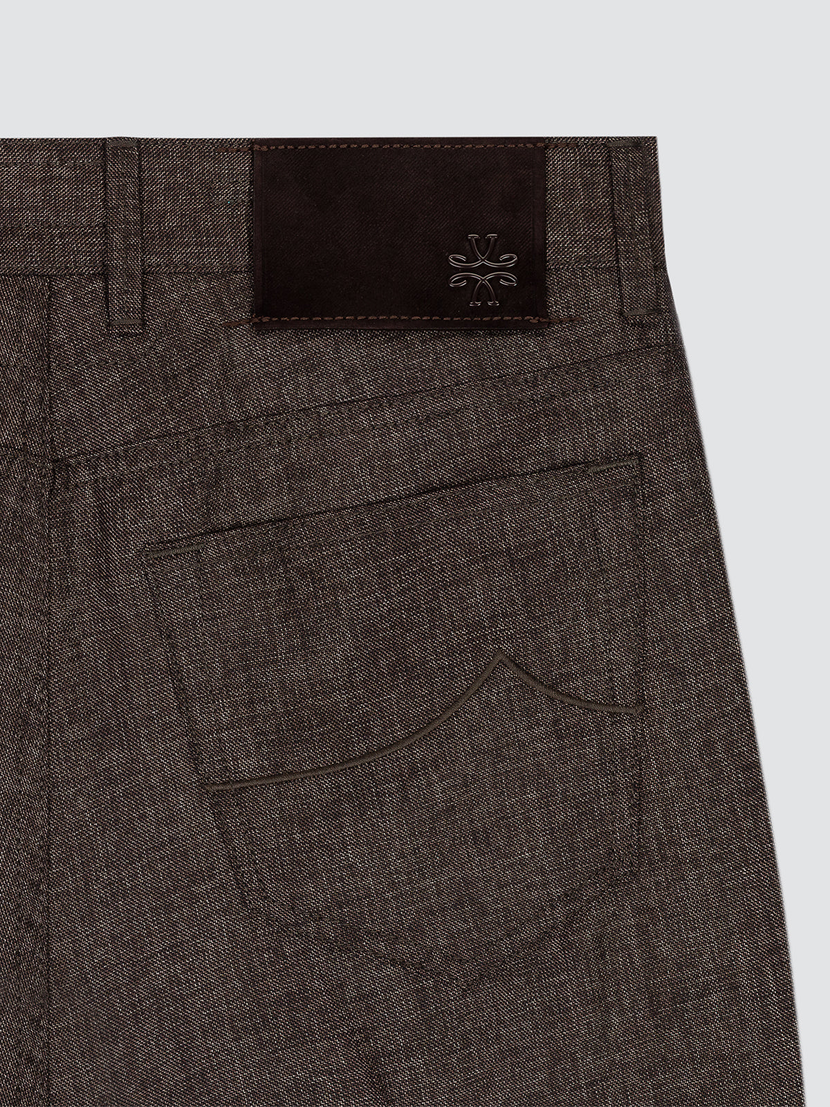 Close-up of the back waistband and pocket area of dark brown textured trousers, featuring a dark brown leather patch with a debossed logo.