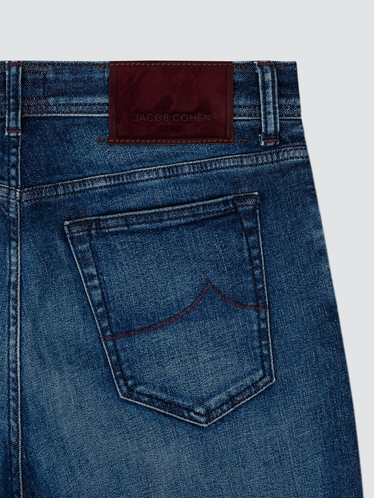 Close-up of the back waistband and pocket of blue denim jeans, showing a dark red leather patch with the embossed brand name JACOB COHEN.