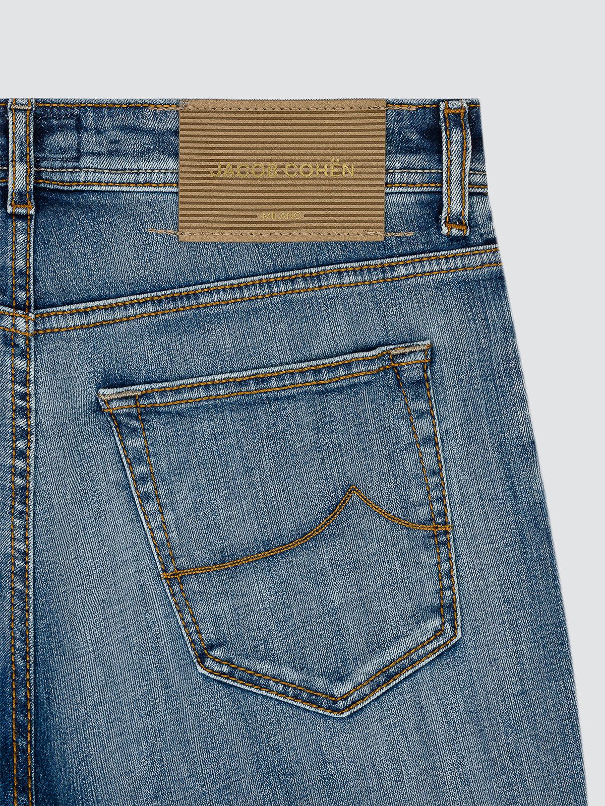 Close-up of the back waistband and pocket of blue denim jeans, showing a tan leather brand patch that reads JACOB COHEN on a light gray background.
