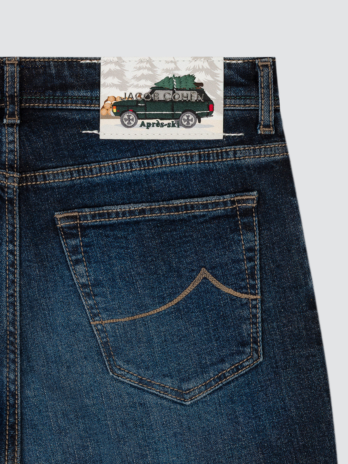 Close-up of the back waistband and pocket of dark blue denim jeans, showing a sewn-on brand patch that reads Jacquetta Anita April-ak.