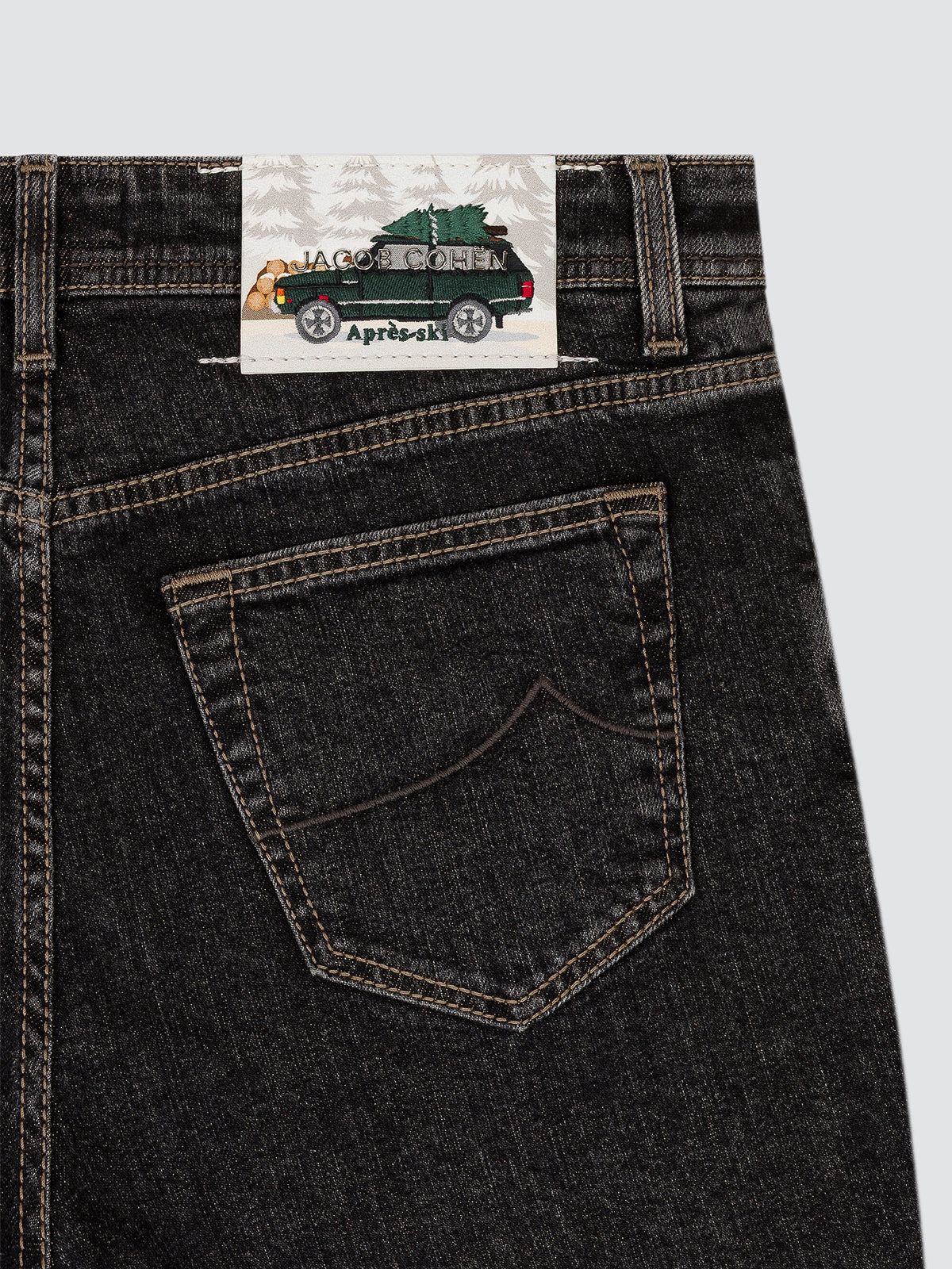 Close-up of the back waistband and pocket of dark washed denim jeans, showing a paper brand label with a green vehicle illustration and the text 'JACOB COHEN.