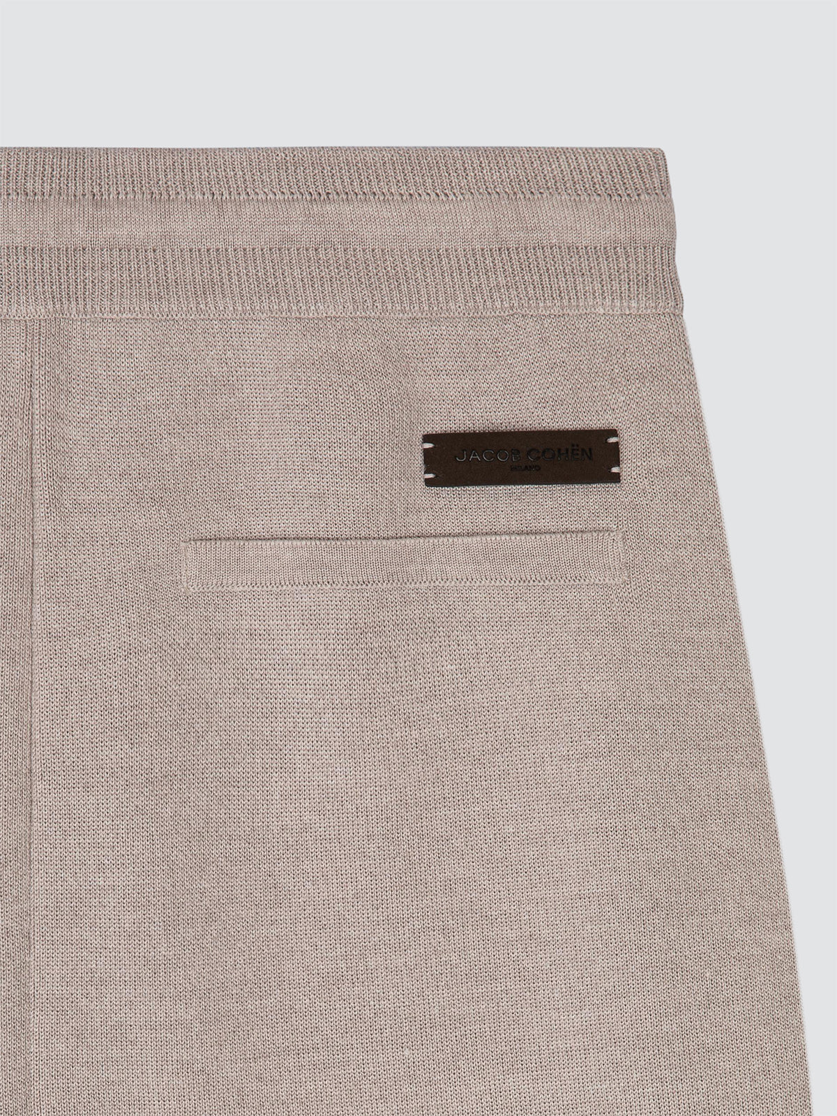 Close-up of a light beige knit garment, likely trousers or a jacket, showing a front welt pocket and a small dark brown rectangular brand plaque on the upper right side.