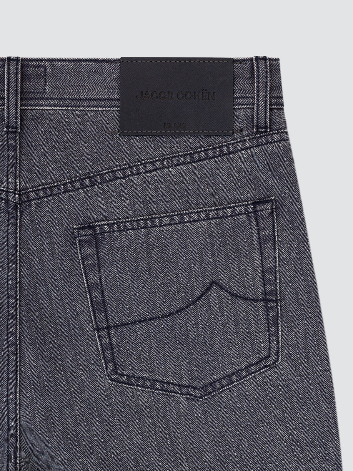 Close-up of the back waistband and rear pocket of dark gray denim jeans, showing a black leather patch with the Jacob Cohën brand name.