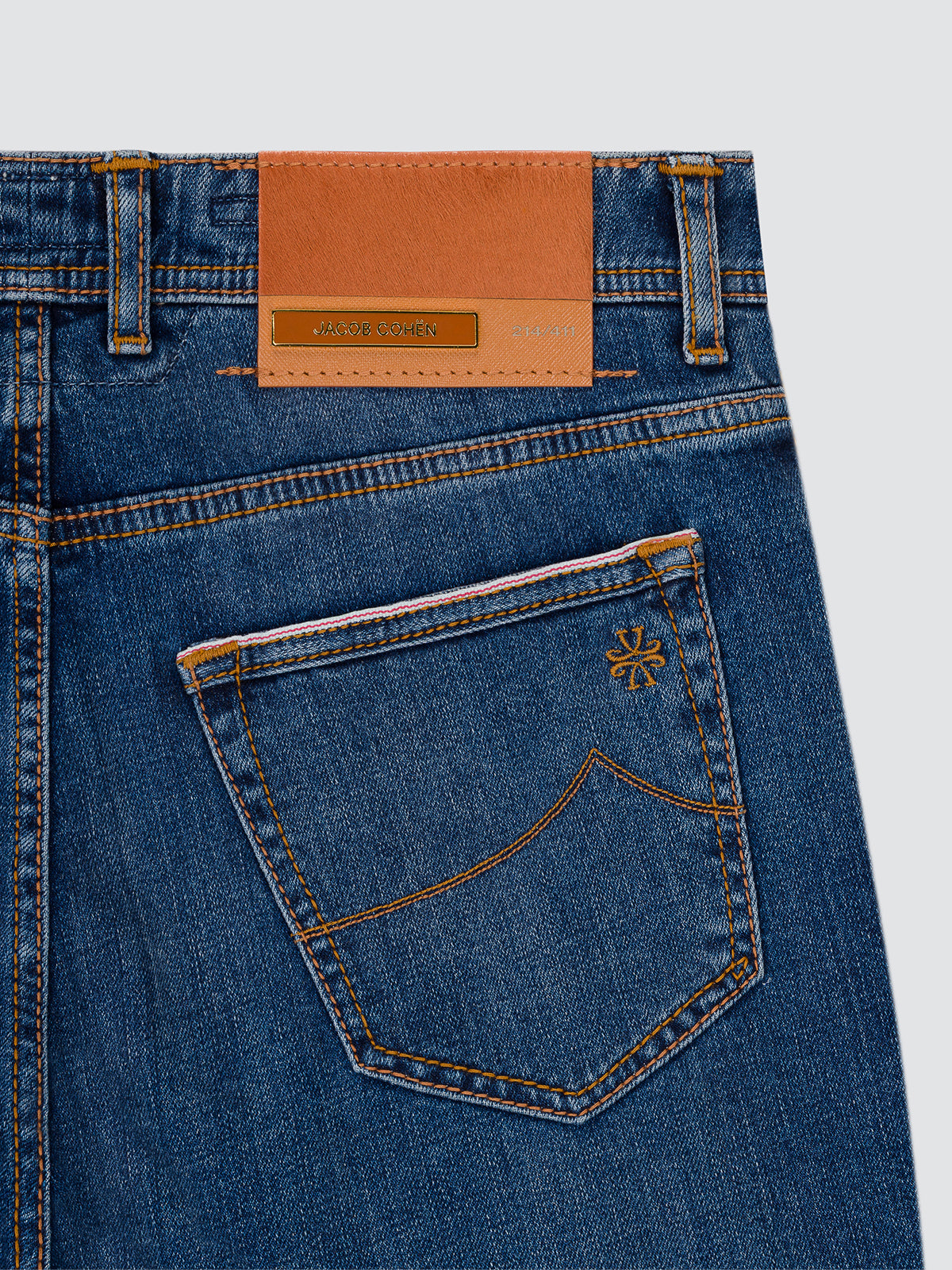 Close-up of the back waistband and pocket area of blue denim jeans, showing a tan leather patch with a metallic logo, contrast stitching, and a small embroidered logo on the pocket. ltd Jacob Cohen bard