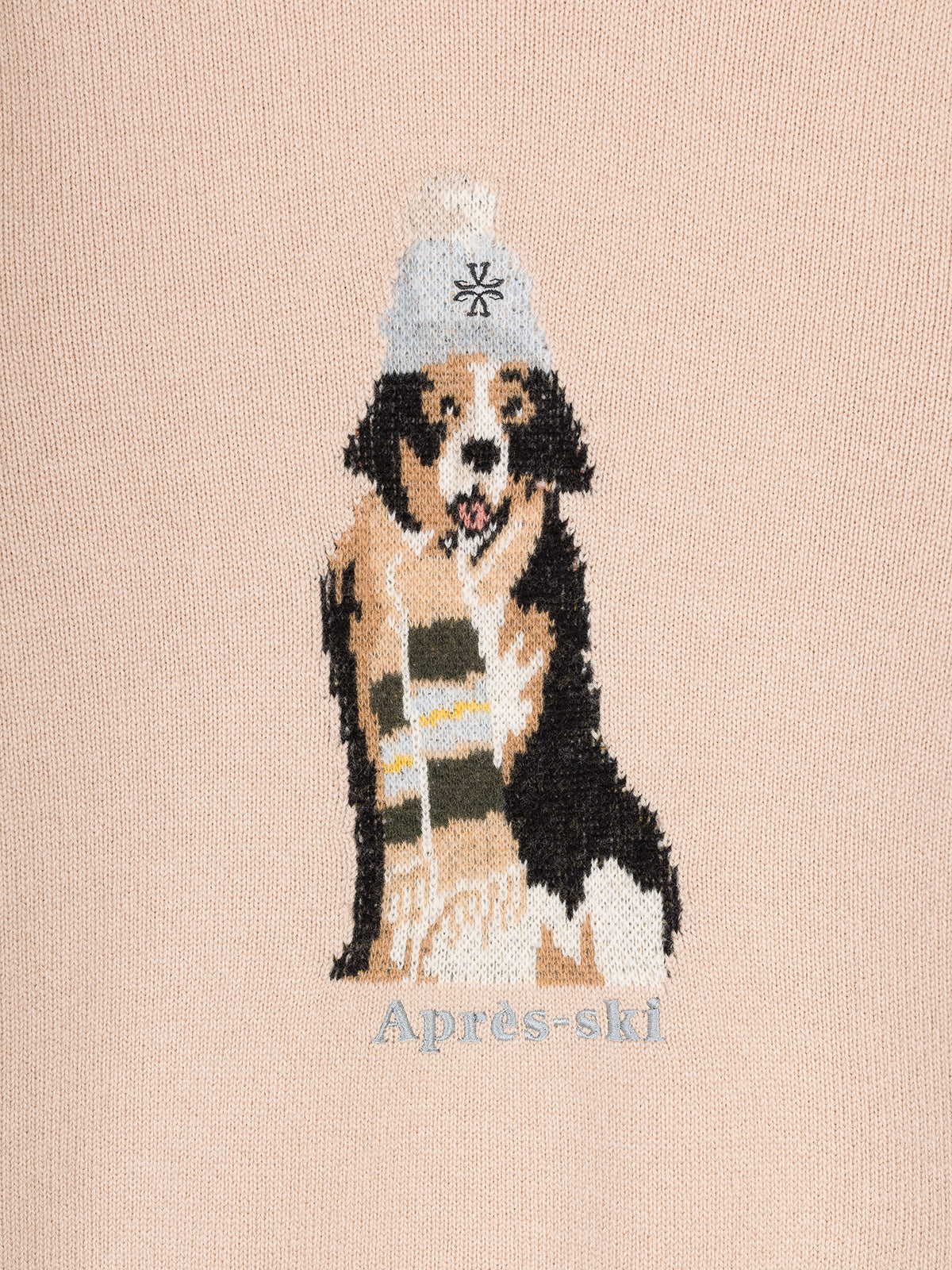 A close-up of a light beige knit fabric featuring a crocheted-style graphic of a dog wearing a white beanie with a black snowflake and a striped scarf, with the text 'Après-ski' below.
