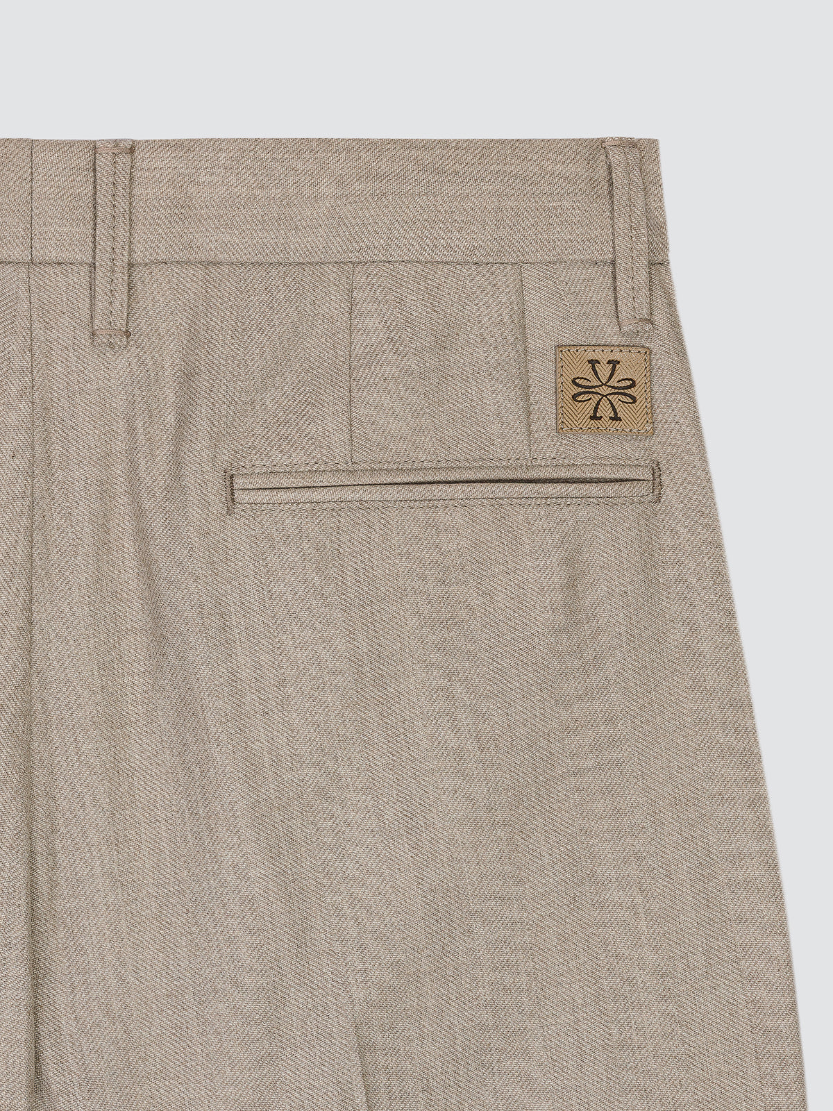 Close-up of the back waistband and pocket area of beige herringbone trousers, featuring a small tan brand patch with a black monogram on the upper right side.
