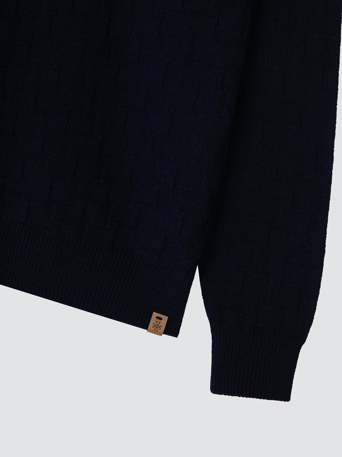 Close-up of a dark navy textured knit sweater sleeve with a ribbed cuff and a small tan brand tag near the hem, shown on a light gray background.