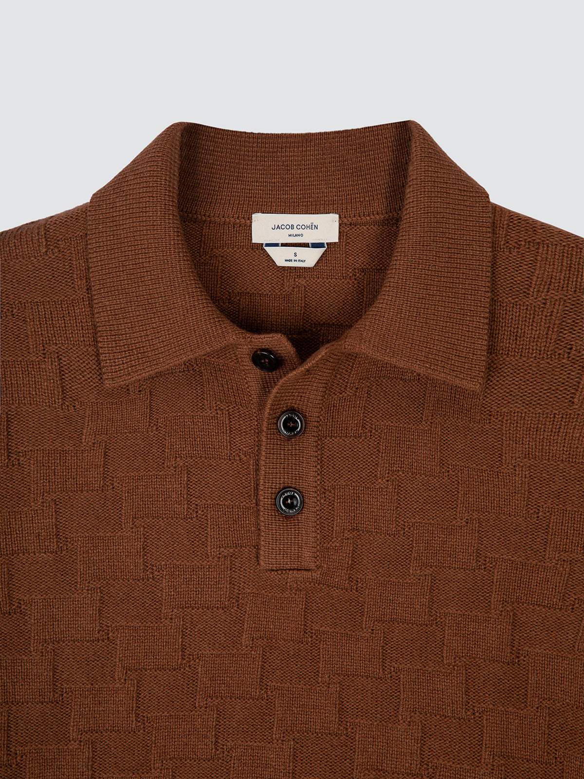 A close-up of a brown textured knit polo shirt by Jacobo Bona, featuring a classic collar, three-button placket, and a subtle basketweave pattern, set against a light gray background.