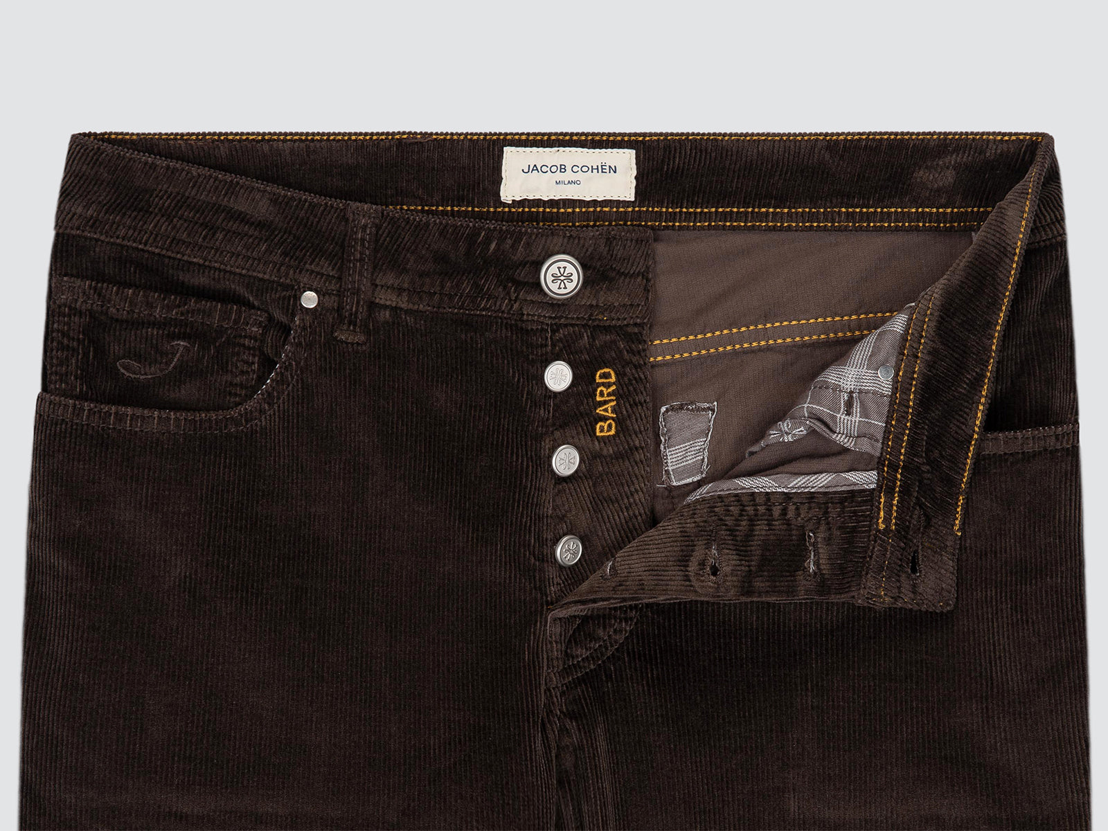 Close-up of dark brown corduroy pants by Jacob Cohen, showing the waistband, button fly, and an opened front pocket revealing a plaid lining. The brand name BARD is embroidered on the side.