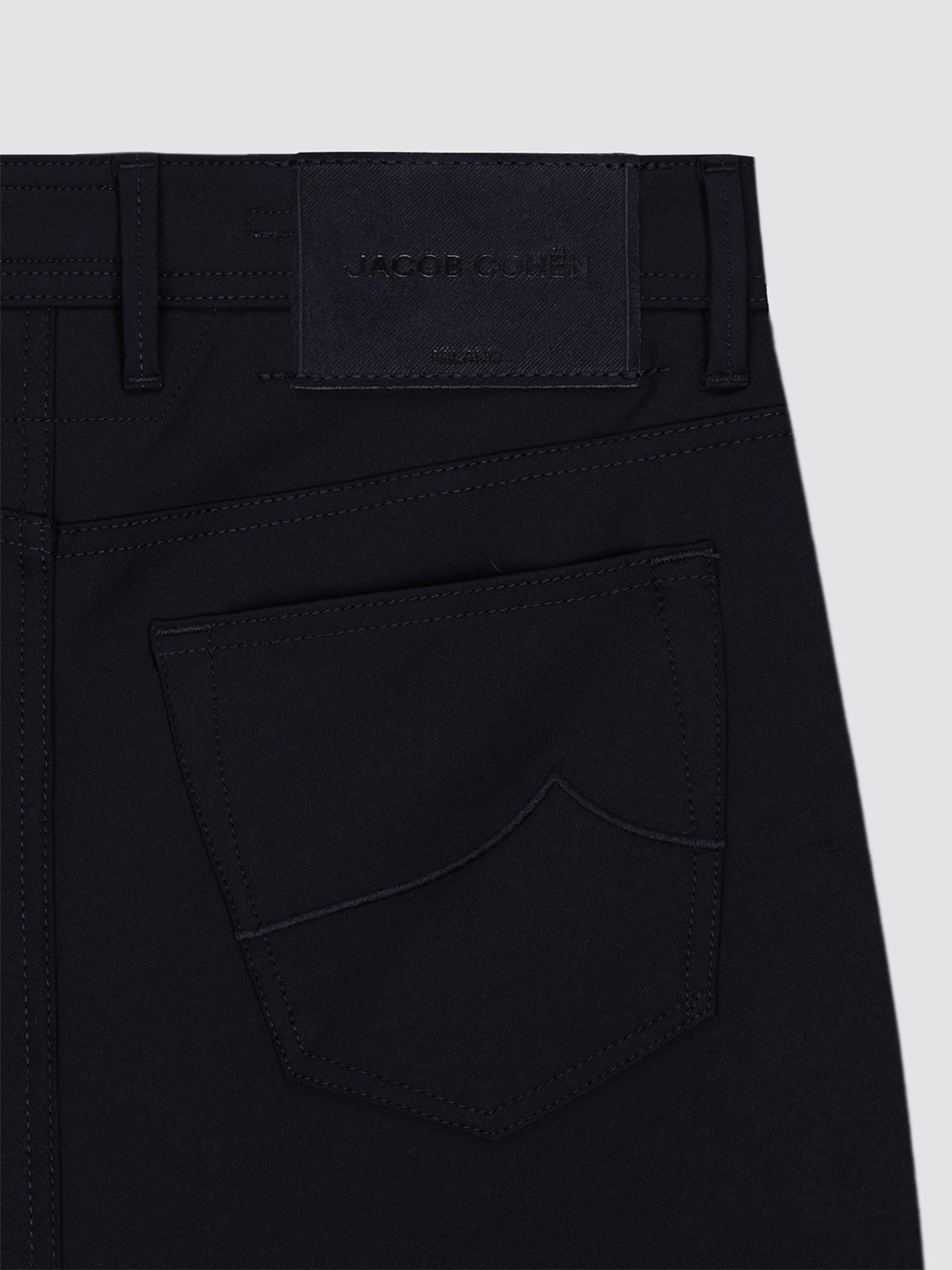 Close-up of the back waistband and pocket area of dark navy tech 5 pockets, showing a branded leather patch at the back waist and tonal stitching on a plain light gray background.