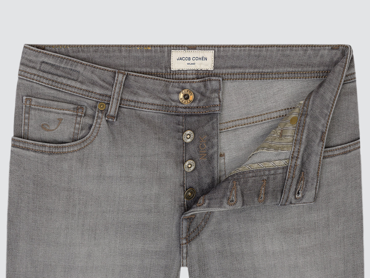 Close-up of gray denim jeans showing the front waistband, button closure, pocket details, and a decorative embroidered patch on the right side. The brand label reads JACOB COHEN.