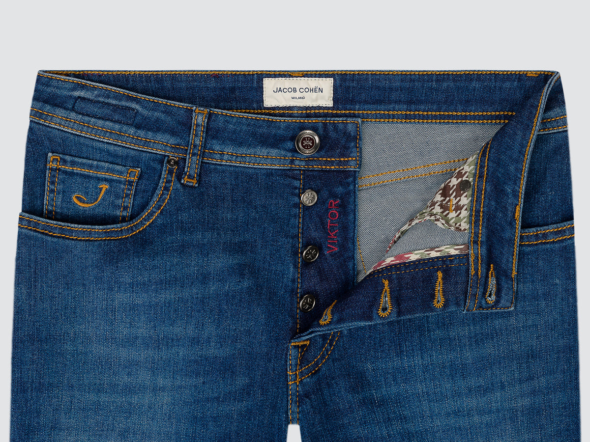 Close-up of a pair of blue denim jeans by Jacob Cohen, showing the front waistband, button closure, embroidered pocket, and a patterned inner lining visible at the opening.