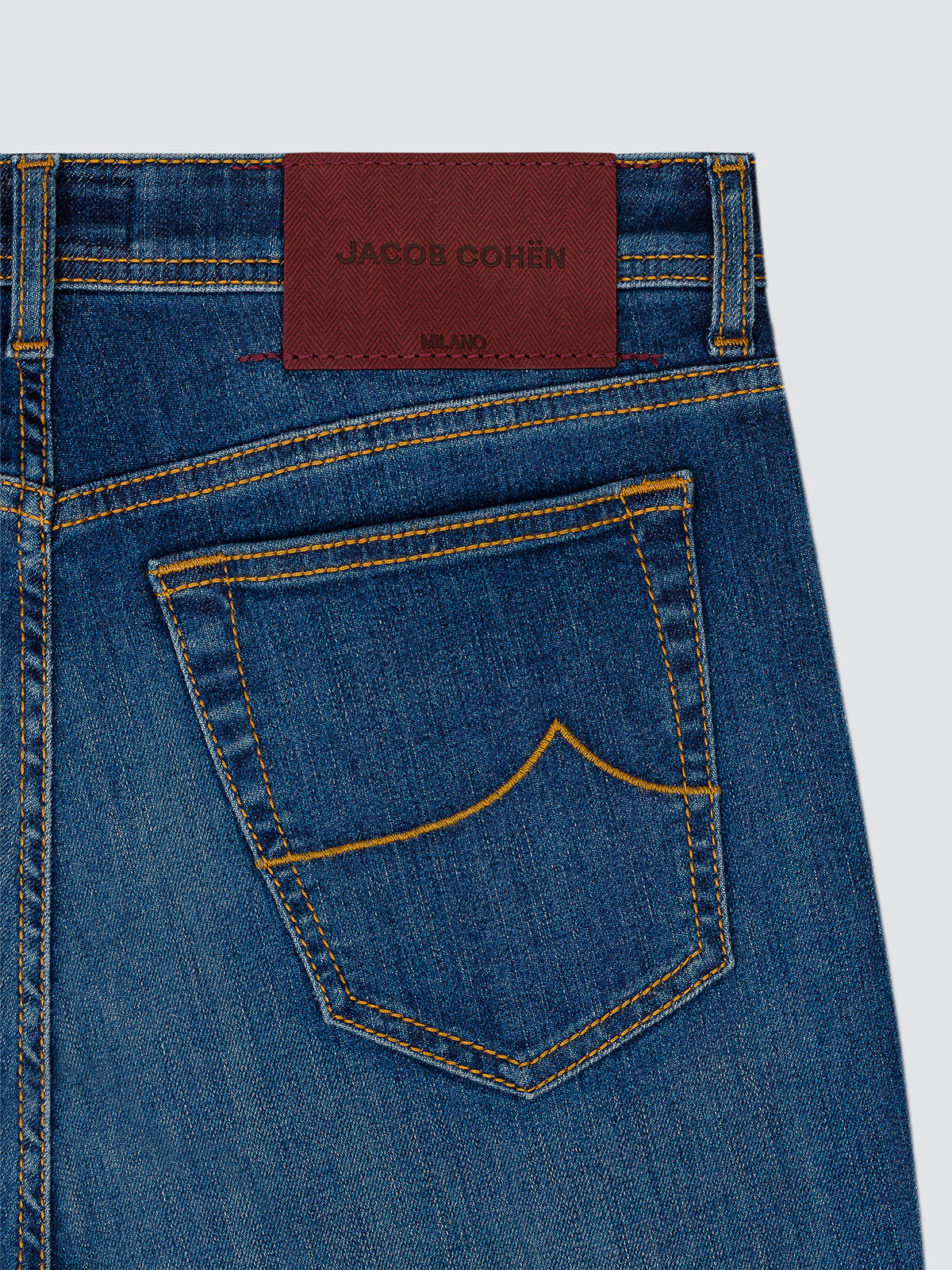 Close-up of the back waistband and pocket of blue denim jeans with a brown leather Jakob Cohen brand patch at the back waist and contrast yellow stitching.