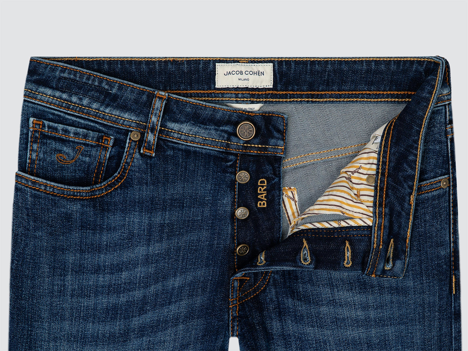 Close-up of a pair of blue denim jeans showing the front waistband, button closure, pocket detail, and an open back pocket revealing a striped lining. The inner waistband label reads JACOB COHEN, and the side embroidery reads BARD.