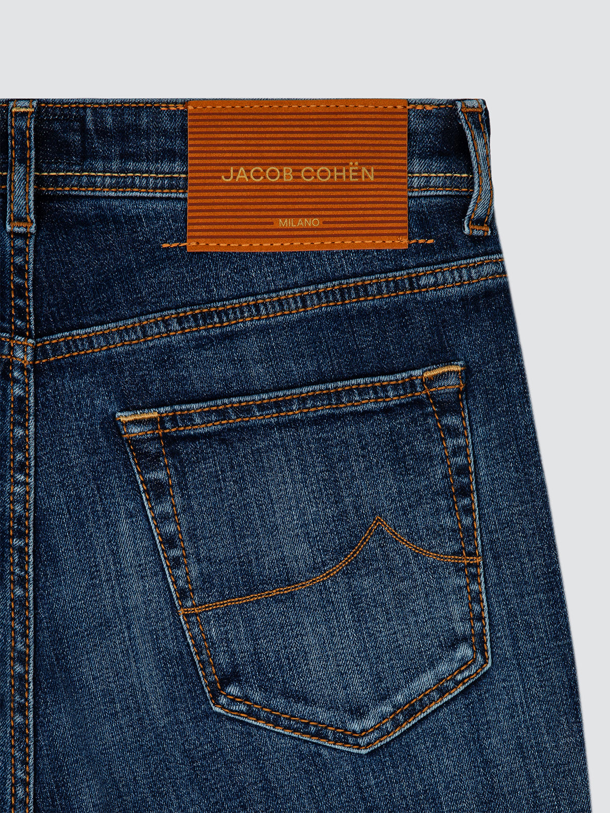 Close-up of the back waistband and pocket of dark blue denim jeans with orange contrast stitching and a visible JACOBE COHËN label.