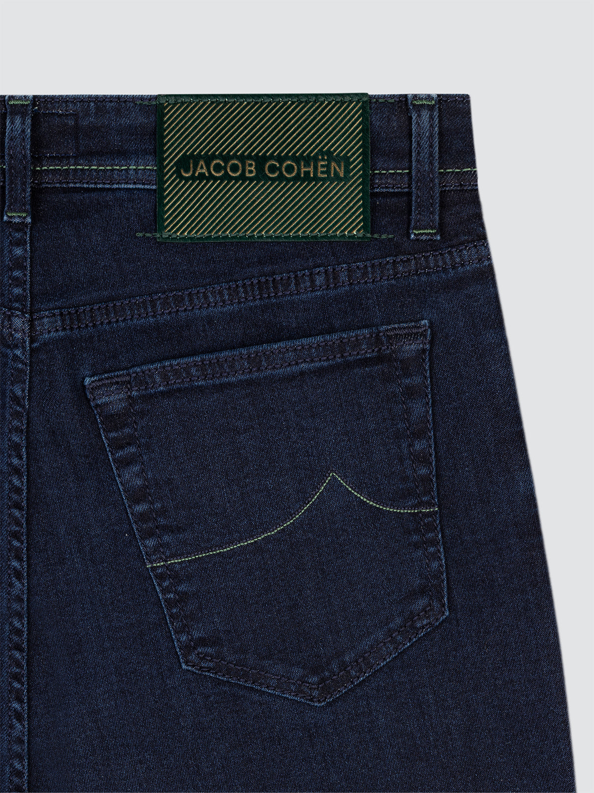 Close-up of dark blue jeans with a green Jacob Cohen label on a light gray background