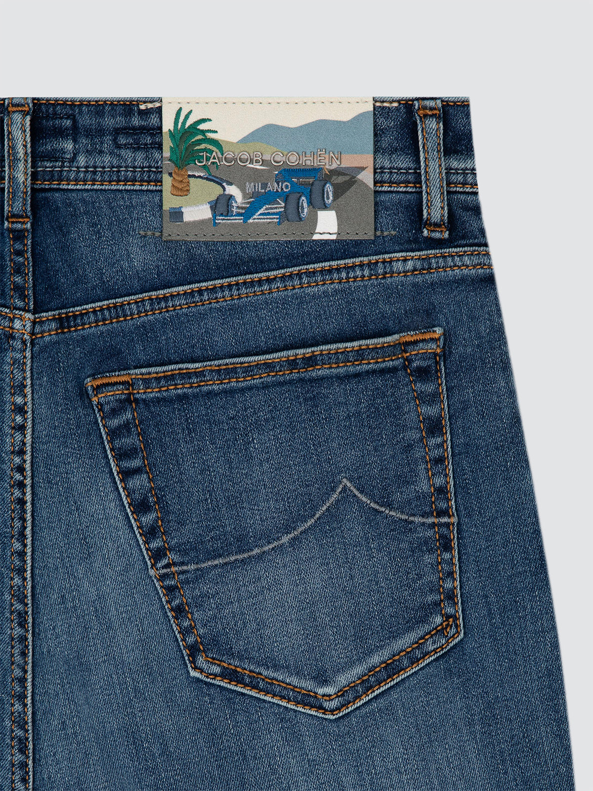 Close-up of the back pocket of blue denim jeans Jacob Cohën with contrast stitching and a decorative brand patch showing a blue race car and palm tree on a light gray background.