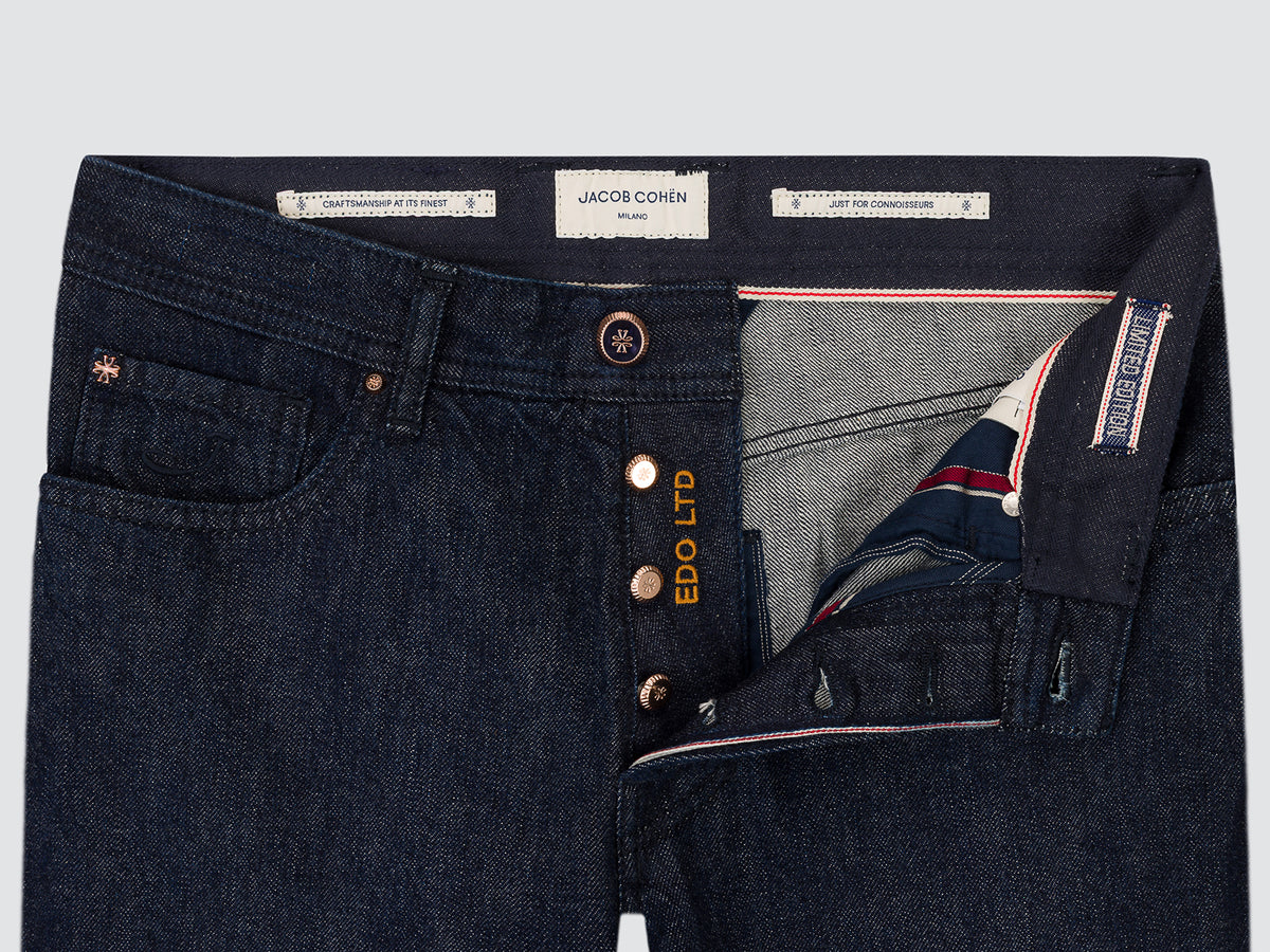 Close-up of dark indigo denim jeans showing the waistband, button closure, interior pocket lining, and interior brand labels including JACOB COHN and EDO LTD.