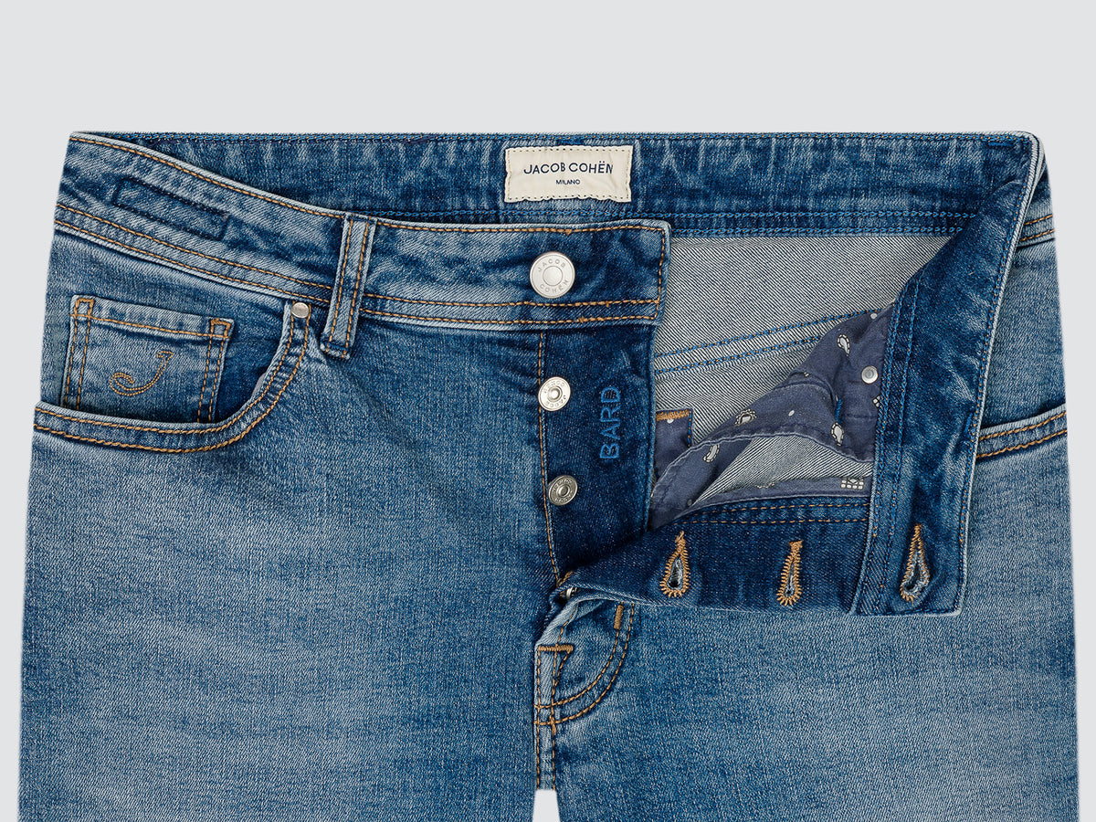 Close-up of blue denim jeans showing the waistband, front button closure, pocket details, and an open back pocket with a patterned lining. A visible waistband label reads Jacob Cohen Milano, and 'Bardo' is printed near the button.