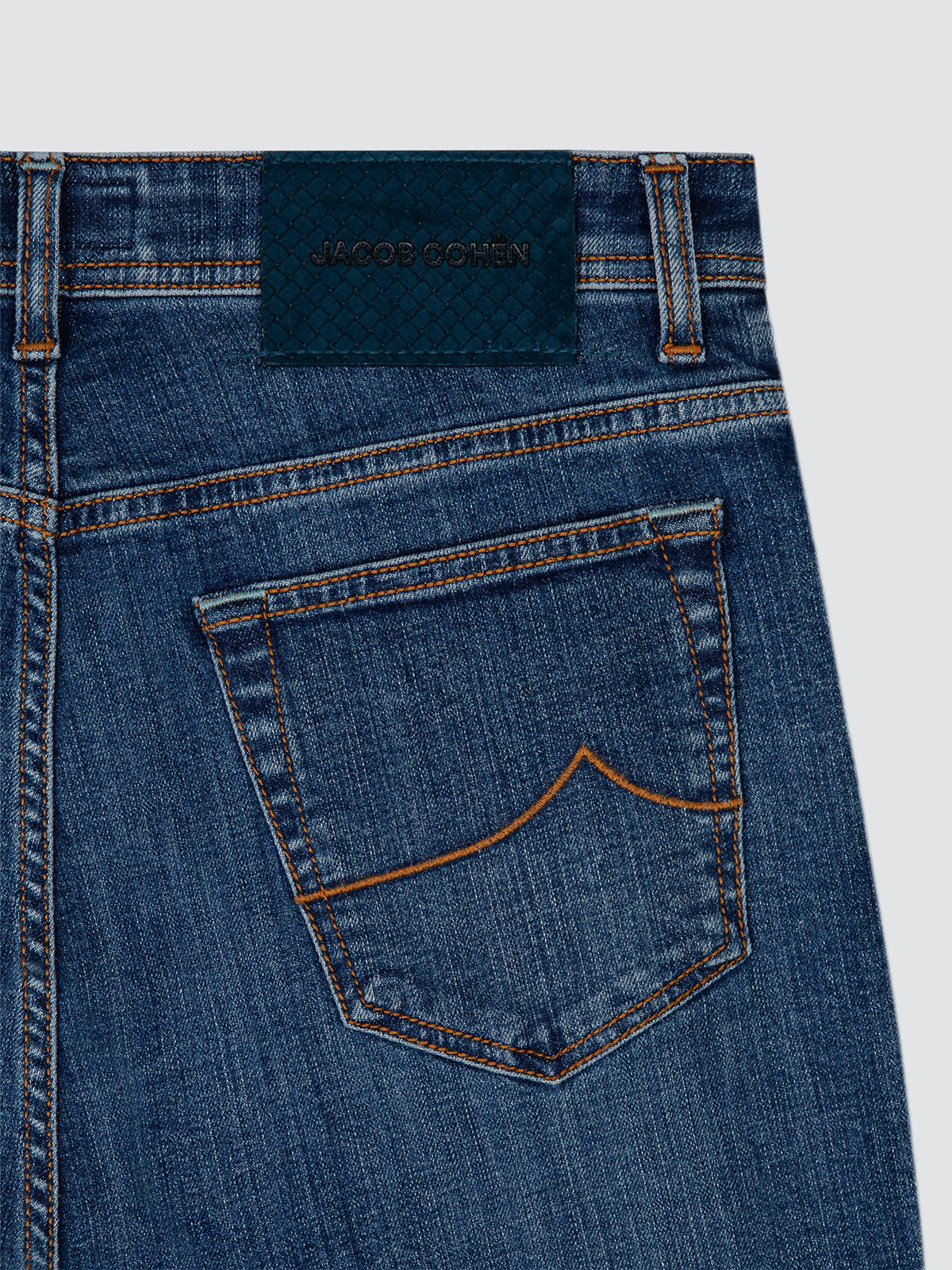 Close-up of the back waistband and pocket of blue denim jeans, showing a dark blue jacquard brand patch with 'JACOB COHEN' embroidered on it, contrast orange stitching, and a light gray background.