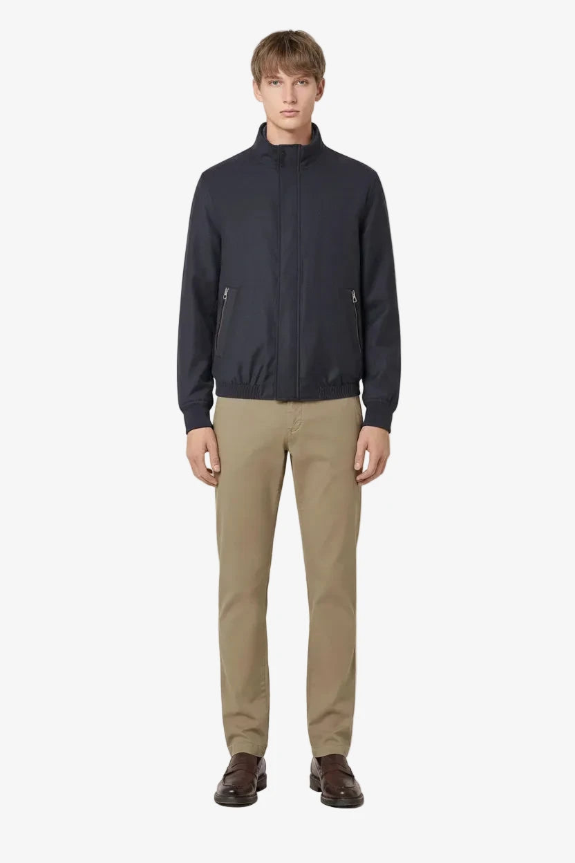 A male model stands against a solid light beige background wearing a dark navy blue zip-up jacket, khaki trousers, and dark brown leather shoes.